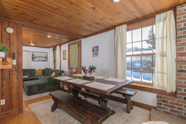 A cozy room with a wooden ceiling, a green sofa, a dining table with placemats, and a large window with a view of a snowy landscape.