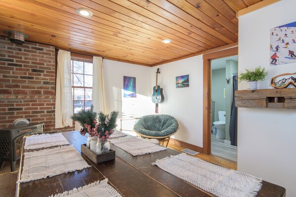 A cozy room with a wooden ceiling, a brick wall, a dining table with placemats, a guitar, and a chair.