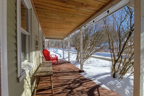 A covered porch with a red chair and a bench, wooden ceiling, and a view of a snowy landscape. - Historic 1880s Cape, 10 Min to Jay Peak | Rustic Charm, Clawfoot Tub & Workspace (Montgomery Center)