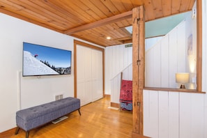Una habitación con techo de madera, un banco, un televisor empotrado mostrando un paisaje nevado de montañas y una escalera con un tapete rojo estampado.