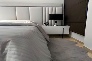 A modern bedroom with a bed, a bedside table with a lamp, and a wall-mounted alarm clock. - The Phoenician Pause (Tunis)