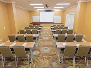 Un salón de conferencias con filas de mesas y sillas, una pantalla de proyector y piso alfombrado.