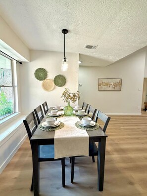 A dining room with a rectangular table set for six, surrounded by chairs, with a vase of flowers on the table. - The Shenandoah Sanctuary: A Fully Furnished Modern Retreat (Richmond)