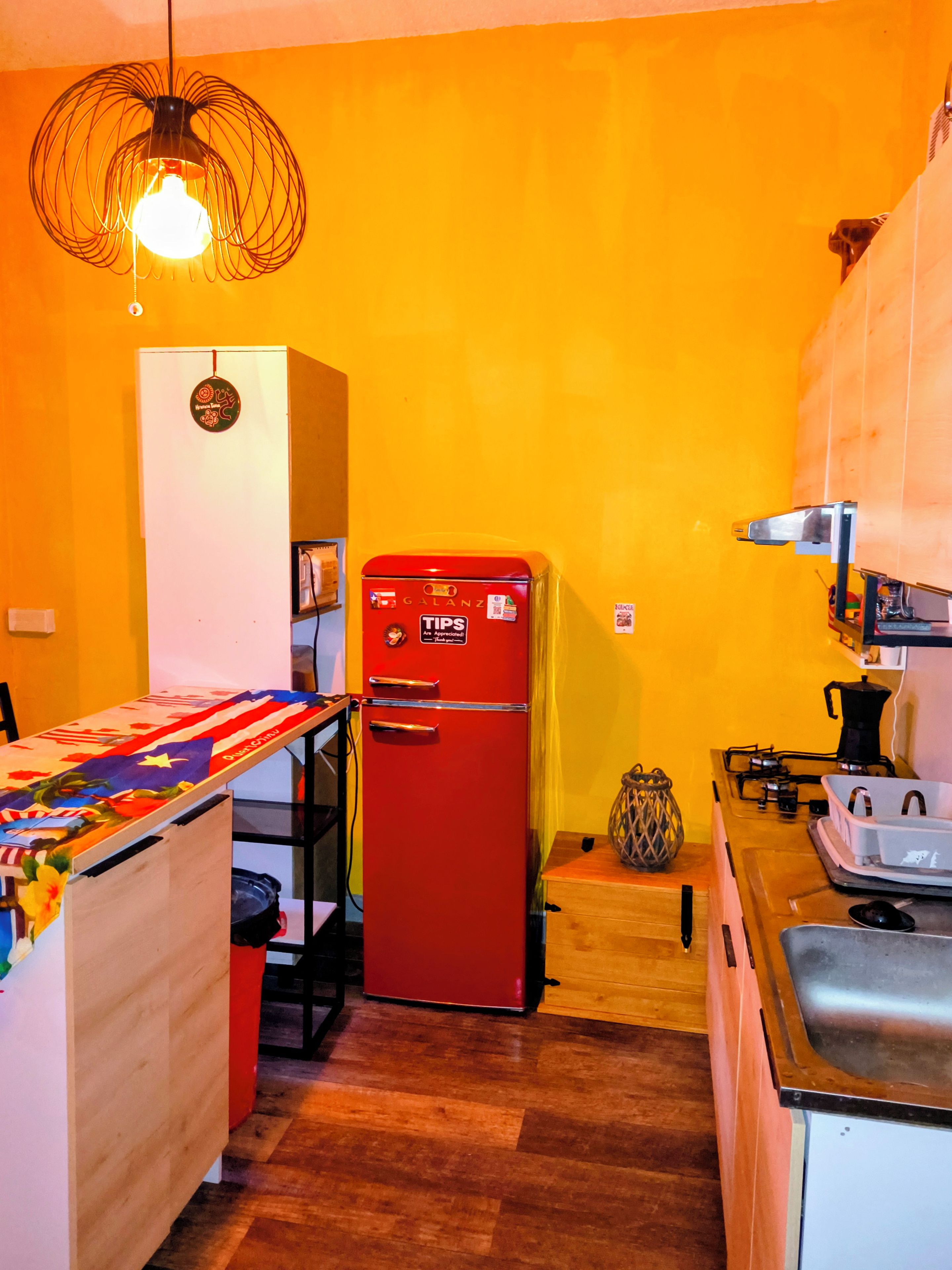 Une cuisine de taille réduite avec un réfrigérateur rouge, une table en bois recouverte d’une nappe colorée et un luminaire suspendu.