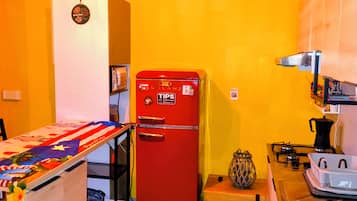Une cuisine de taille réduite avec un réfrigérateur rouge, une table en bois recouverte d’une nappe colorée et un luminaire suspendu.