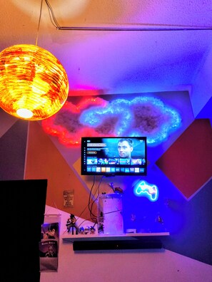 A room with a television mounted on the wall, a neon light, and a shelf with various items.