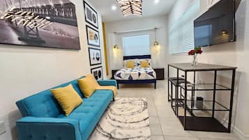 Uma sala de estar moderna com um sofá azul, uma cama com roupa de cama estampada, uma mesa de centro e uma TV fixada na parede.