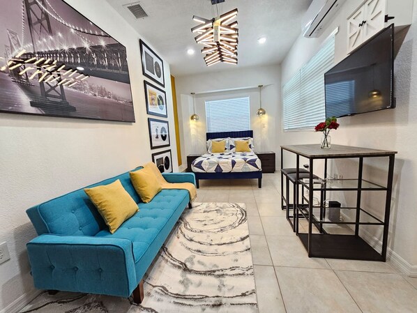 Uma sala de estar moderna com um sofá azul, uma cama com roupa de cama estampada, uma mesa de centro e uma TV fixada na parede.