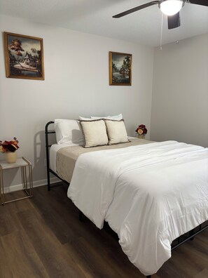 Una camera da letto con un letto, due quadri appesi al muro, una ventola a soffitto e un comodino con un vaso di fiori.