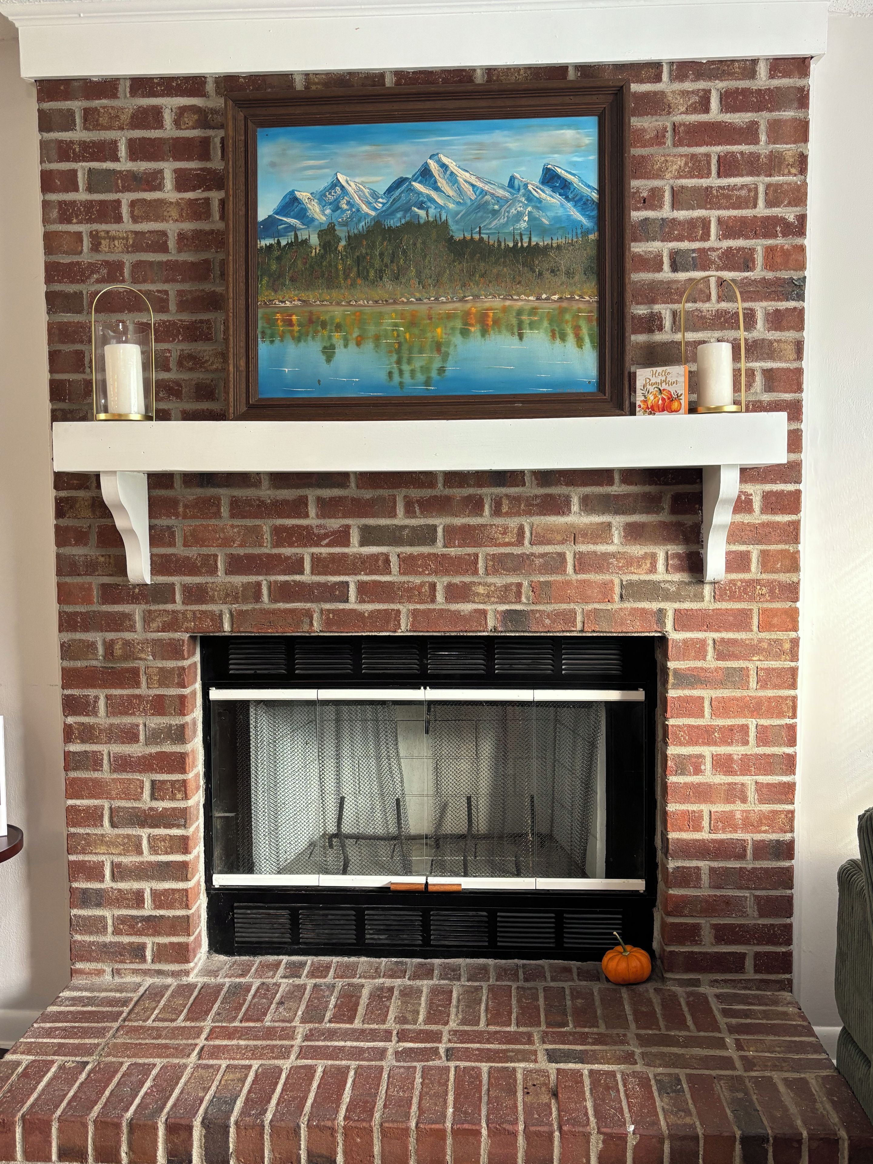Een metselwerk open haard met daarboven een lijstwerk schilderij van een landschap, een witte schoorsteenmantel met decoratieve objecten, en een zwarte glazen deur.