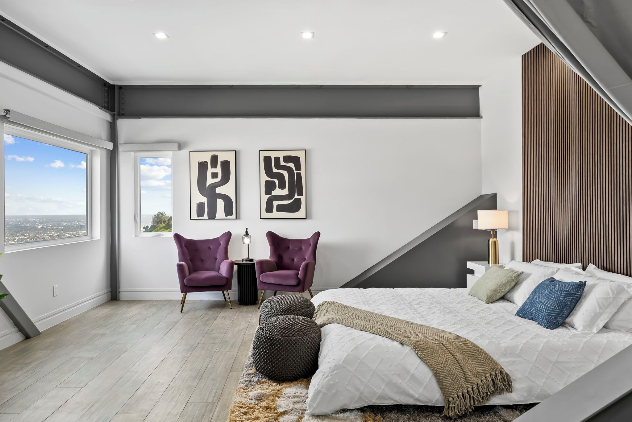 A modern bedroom with a large bed, two chairs, a view of the city, and abstract art on the wall.