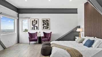 A modern bedroom with a large bed, two chairs, a view of the city, and abstract art on the wall.