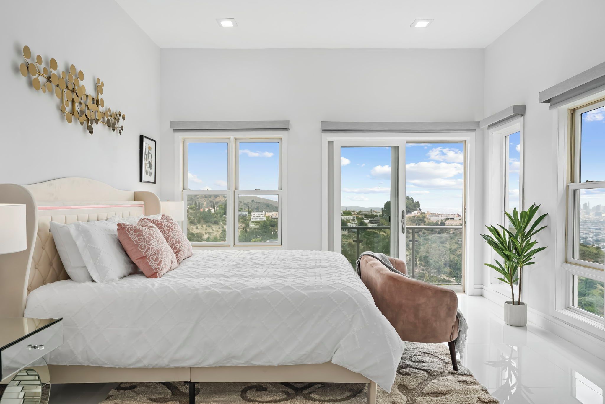 A bedroom with a large bed, a nightstand, a chair, a potted plant, and a view of a cityscape through large windows.