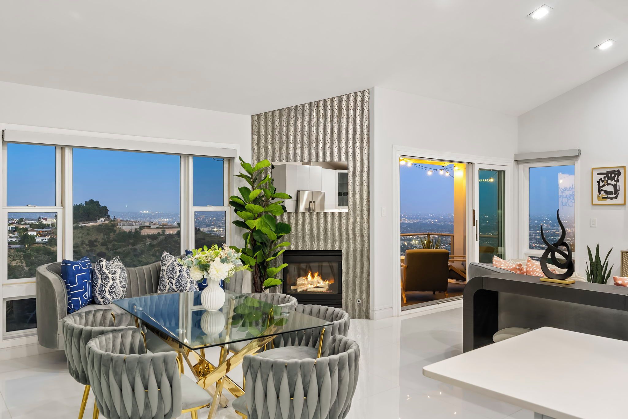 A modern living room with a glass-top dining table, grey upholstered chairs, a fireplace, and large windows offering a city view.