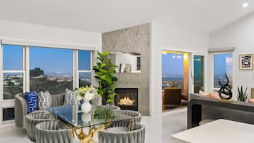 A modern living room with a glass-top dining table, grey upholstered chairs, a fireplace, and large windows offering a city view.