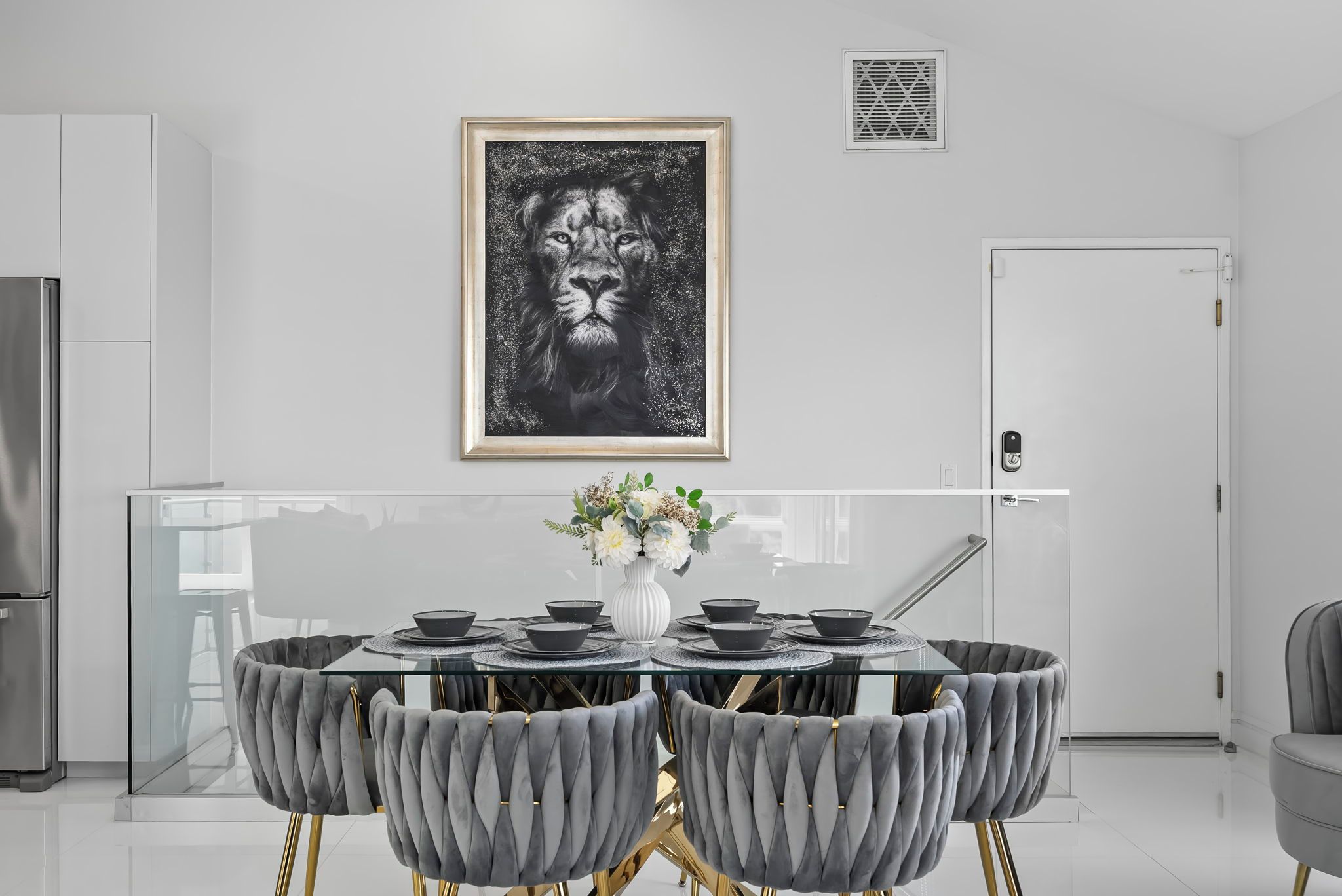 A modern kitchen with a dining area featuring a glass-top table, grey upholstered chairs, and a framed artwork above.