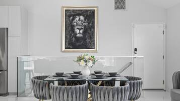 A modern kitchen with a dining area featuring a glass-top table, grey upholstered chairs, and a framed artwork above.