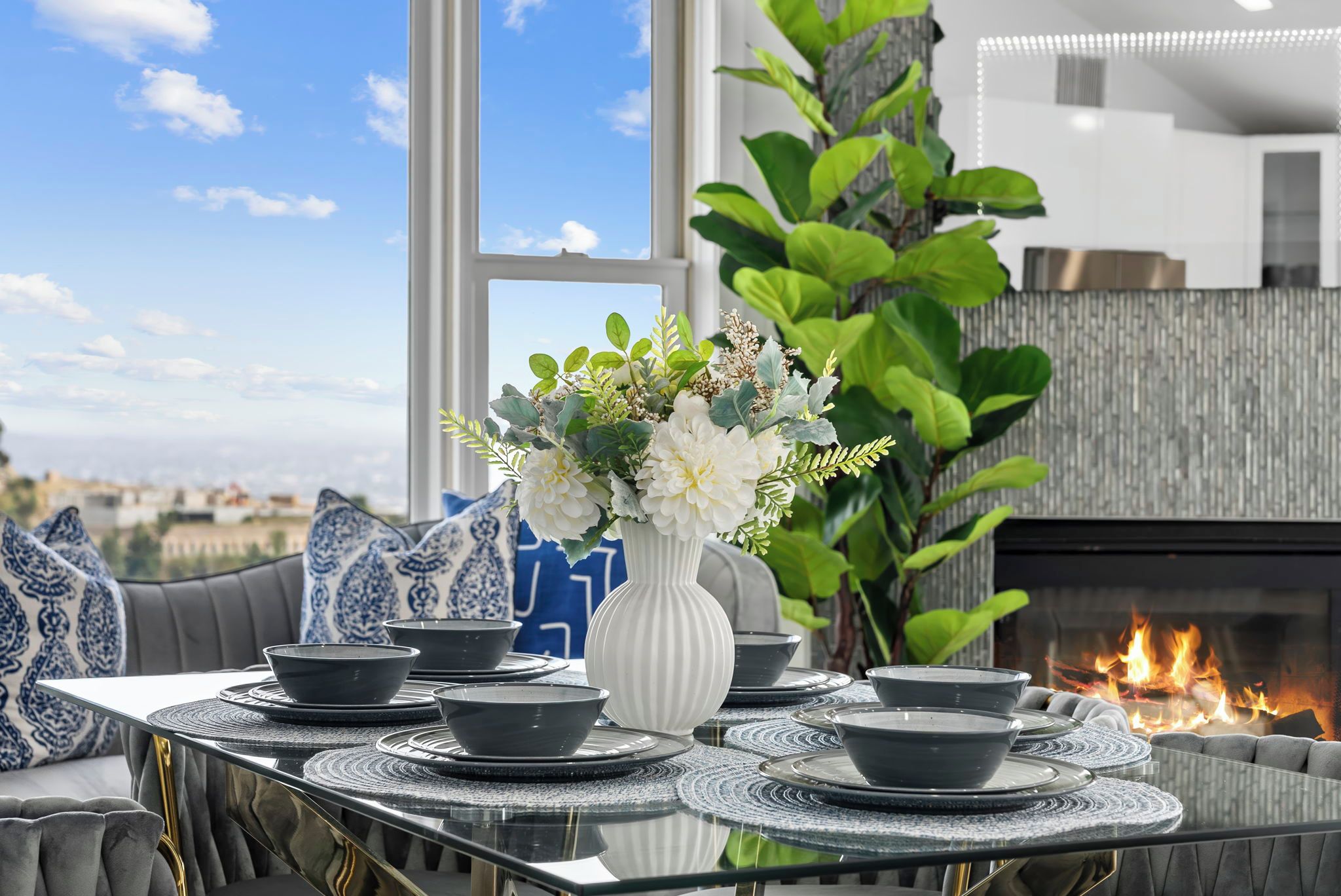 A dining area with a table set for four, a white vase with flowers, and a fire feature in the background.
