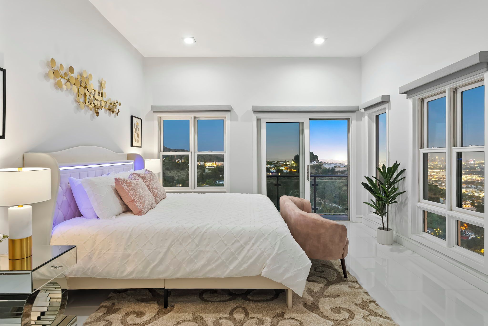 A modern bedroom with a large bed, a bedside table, a lamp, a potted plant, and a view of a cityscape through the windows.