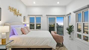 A modern bedroom with a large bed, a bedside table, a lamp, a potted plant, and a view of a cityscape through the windows.