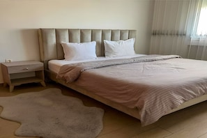 Un dormitorio con cama, mesita de noche, alfombra grande y ventana con cortinas traslúcidas.