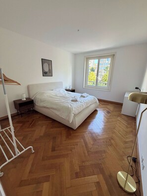 A hotel room with a large bed, a bedside table, a nightstand, a window with a view, a floor lamp, and a wall-mounted picture. - Spacious 2-Bedroom Apartment in Central Lausanne (Lausanne)