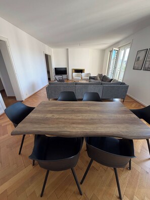 Een moderne woonkamer met een grote houten eettafel, zwarte stoelen, een grijze bank en een flatscreen televisie.