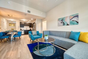 A modern living room with a grey sofa, blue chairs, a round coffee table, and a dining area with a table set for two. - Vista Cay 3BR Condo King Suite Near Universal (Orlando)