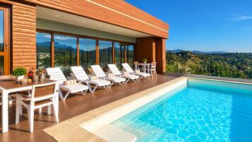 Un área de piscina moderna al aire libre con sillones de descanso y una mesa, rodeada de una terraza de madera y ventanales.