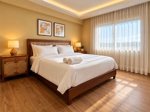 A hotel room with a large bed, wooden headboard, two bedside tables with lamps, a large window with sheer curtains, and two framed artworks on the wall.
