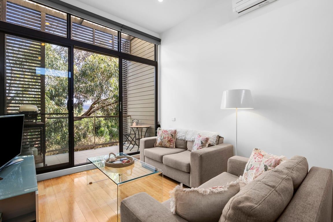 A modern living room with a glass coffee table, two sofas, and a large window with blinds.