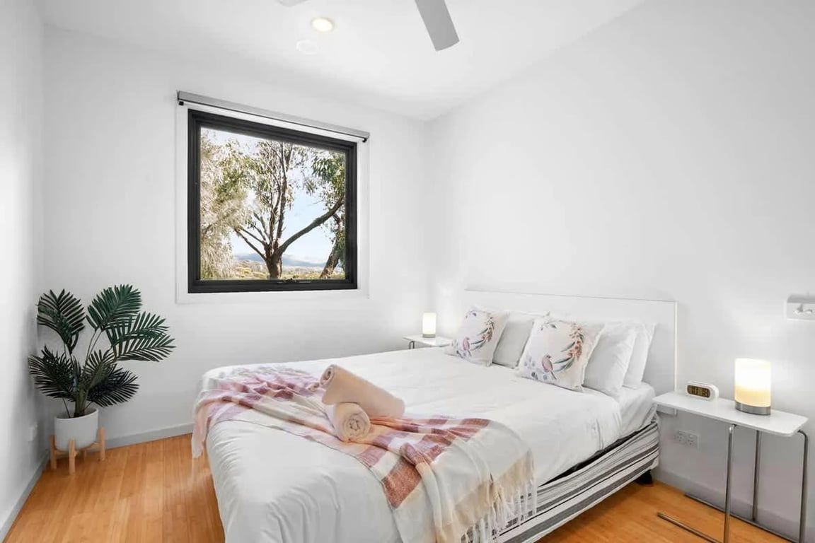 A bedroom with a bed, bedside tables, a nightstand, a potted plant, and a window with a view of trees.