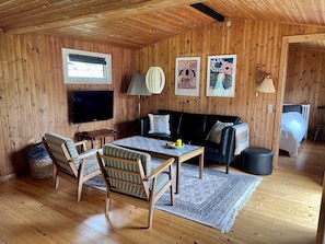 A wooden-paneled room with a sofa, chairs, a table, a TV, and artwork on the walls. - Beautiful cottage close to forest and beach (Væggerløse)
