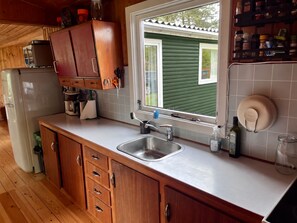A kitchen with wooden cabinets, a stainless steel sink, and a window with a view of a green house. - Beautiful cottage close to forest and beach (Væggerløse)