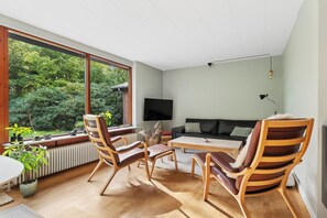 A modern living room with a large window, a flat-screen TV, a black leather sofa, and wooden chairs. - Holiday home near lake, nature and coast (Faldsled)