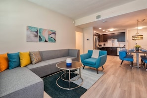Ein modernes Wohnzimmer mit einem grauen Ecksofa, einem runden Couchtisch und einem blauen Sessel. Im Hintergrund ist eine Küche mit dunklen Schränken und Edelstahlgeräten zu sehen.