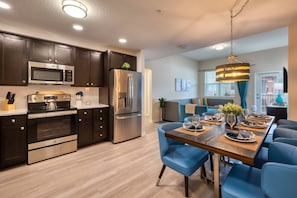 A modern kitchen with stainless steel appliances, a dining area with blue chairs, and a living room with a sofa and a large window. - Vista Cay 3BR Condo King Suite Near Universal (Orlando)