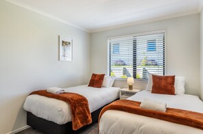 Een hotelkamer met twee bedden, een raam met luxaflex, een nachtkastje met een lamp en een lijst met een afbeelding aan de muur.