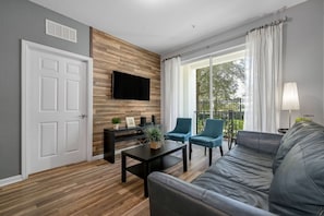 Una sala de estar moderna con un televisor de pantalla plana montado en una pared de madera, una mesa de centro negra, dos sillones azules, un sofá gris y un balcón con cortinas blancas.