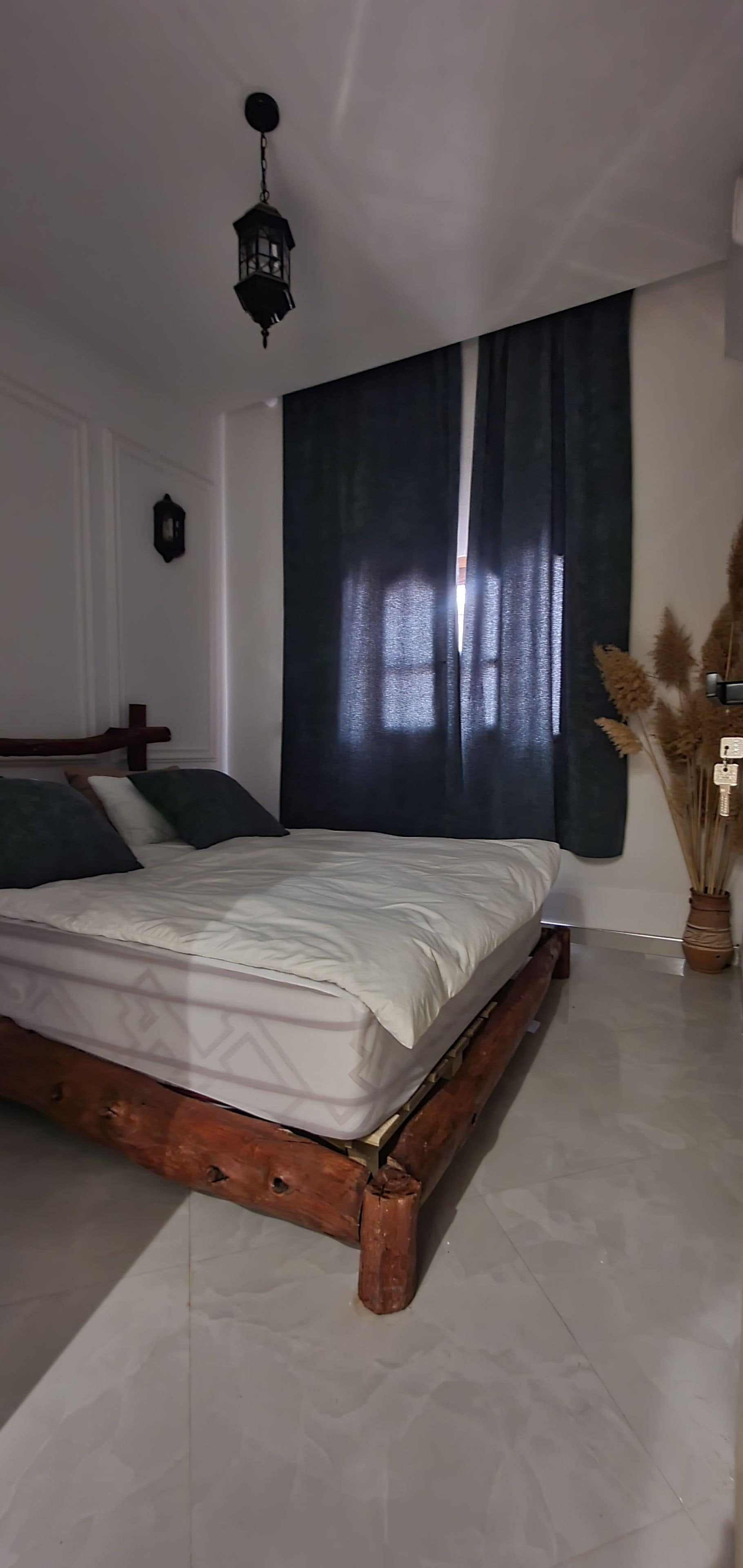 Une chambre à coucher avec un cadre de lit en bois, une grande fenêtre avec des rideaux foncés et un luminaire suspendu.