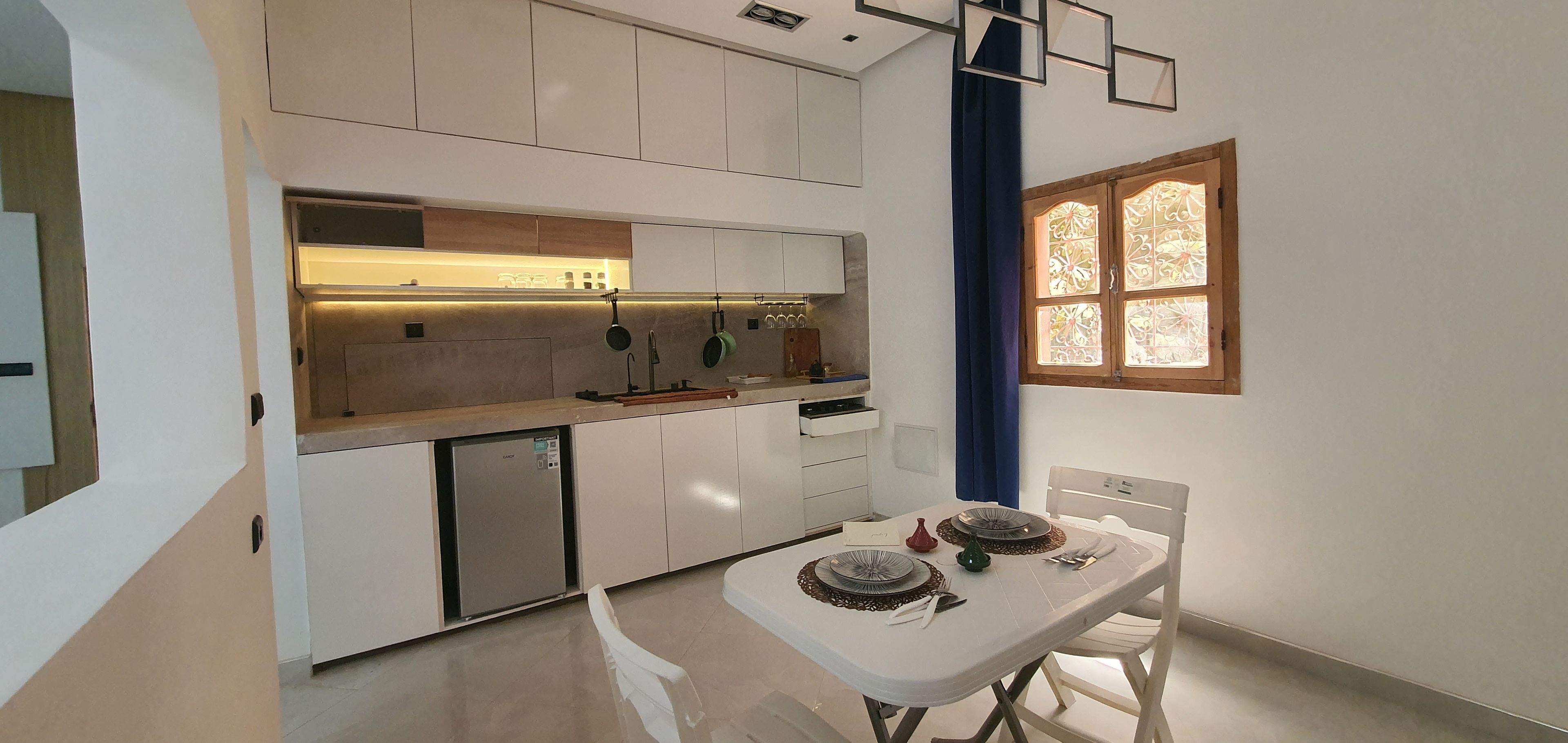 Une cuisine compacte dotée d’appareils électroménagers intégrés, un coin repas avec une table pour deux personnes et une fenêtre ornée de vitraux décoratifs.