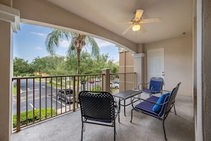 A balcony with a ceiling fan, two chairs with cushions, a small table, and a view of a street with cars and trees. - Vista Cay 3BR Condo Pools Near Universal (Orlando)