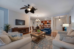 A living room with a ceiling fan, a flat-screen TV, a dining area with a chandelier, and a kitchen with wooden cabinets. - Vista Cay 3BR Condo Pools Near Universal (Orlando)