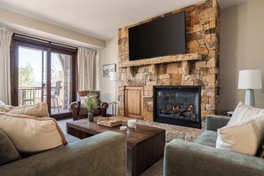 A living room with a stone fireplace, a flat-screen TV mounted above it, and a balcony with a view. - Walk to Frostwood Gondola, private hot tub (Park City)
