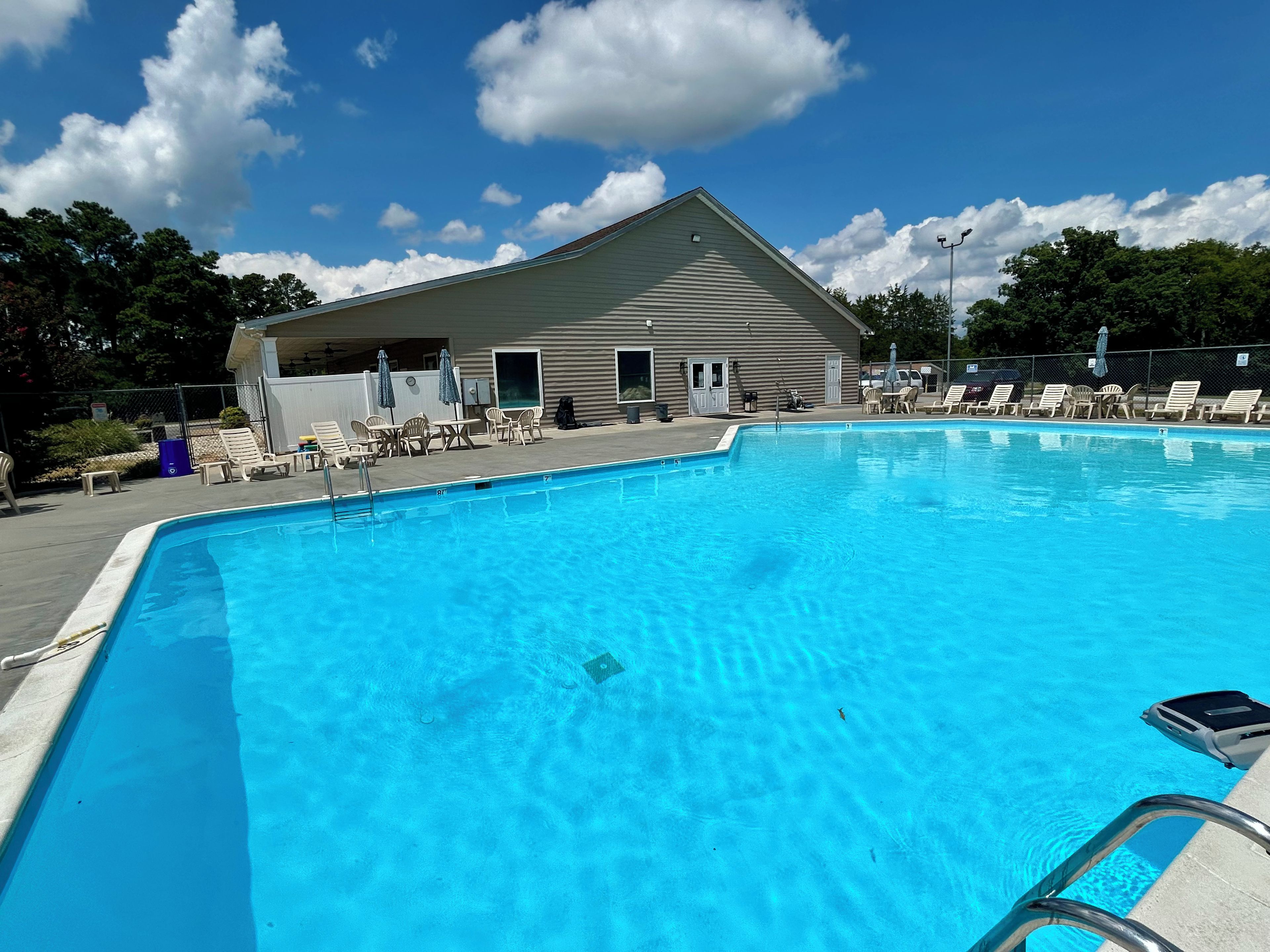 Una piscina con sillones de descanso y sombrillas, un edificio con tejado inclinado y un cielo azul despejado.