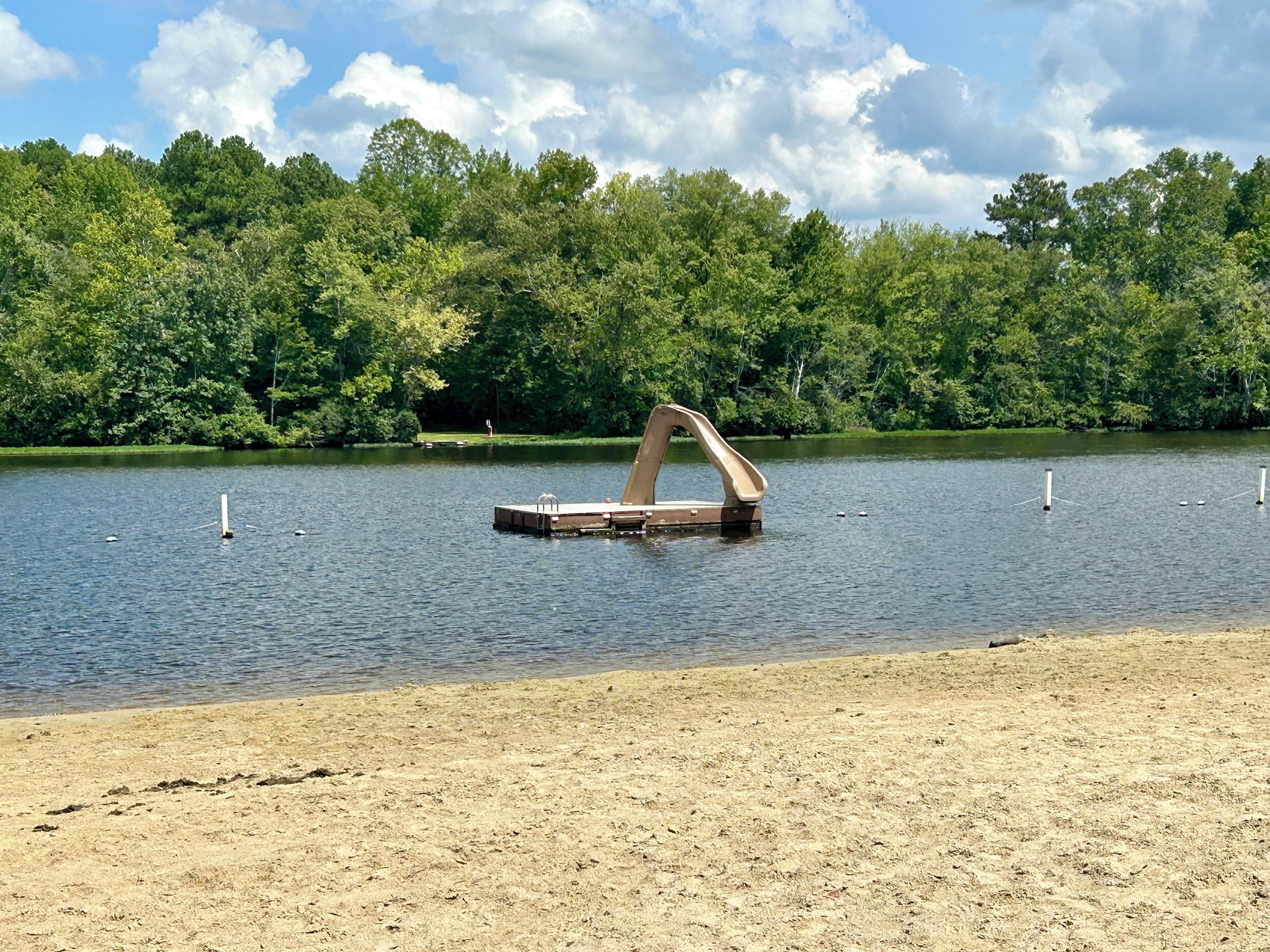 A sandy beach with a wooden boat structure in the water, surrounded by dense green trees and a partly cloudy sky.