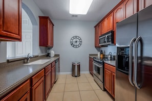A kitchen with wooden cabinets, a stainless steel refrigerator, a microwave, and a clock on the wall. - Vista Cay 3BR Condo King Suite Near Universal (Orlando)