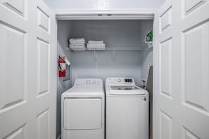A laundry room with a front-loading washer and dryer, a fire extinguisher, and a shelf with folded towels. - Vista Cay 3BR Condo King Suite Near Universal (Orlando)