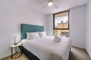 A hotel room with a bed, a bedside table, a ceiling fan, and a window with a view of buildings. - Chic 1 Bedroom Apartment in a Prime Location (Fortitude Valley)