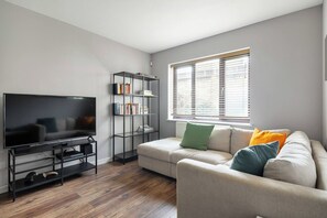 A living room with a flat-screen TV, a bookshelf, a sofa with cushions, and a window with blinds. - Charming Parkside Apartment | WiFi | Gated Parking (Greater London)